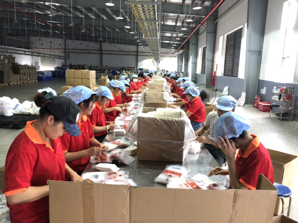Nhiều công nhân mặc áo đỏ đóng gói hàng vào thùng carton trong nhà xưởng lớn